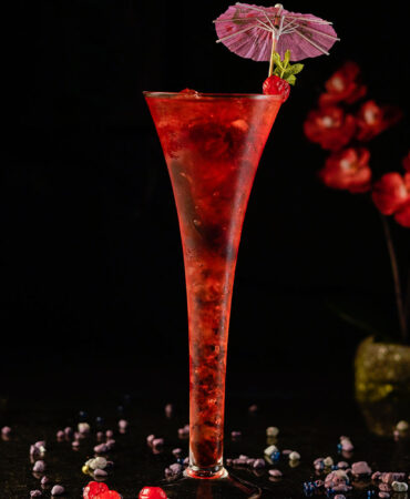 Red cocktail inside a long trumpet champagne glass garnished with an umbrella cocktail pick