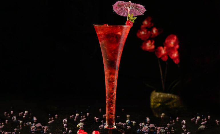 Red cocktail inside a long trumpet champagne glass garnished with an umbrella cocktail pick