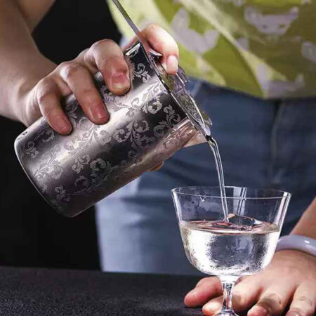 Cocktail being strained from an engraved stainless steel tin into a Glass