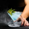 Hand holding The Holy Cone Glass with blue liquid, ice cubes, and mint leaf, surrounded by vapor.