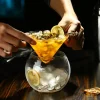 lose-up of hands preparing a lemon cocktail in The Holy Cone Glass with ice and lemon slices.