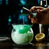 Hand pouring liquid into The Holy Cone Glass with blue-green cocktail, ice, and lemon.