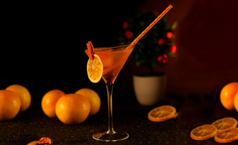 Orange cocktail in a martini glass placed in a dark background next to oranges