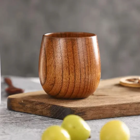 A handcrafted wooden cup from The Artisan Sip, placed on a wooden board with green grapes in the foreground and a blurred gray background, showcasing the natural wood grain and artisanal quality.