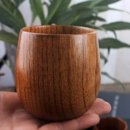 A handcrafted wooden cup with a handle from The Artisan Sip, held in a hand, showcasing the smooth, rounded shape and natural wood grain, with another smaller cup and a green plant in the background.