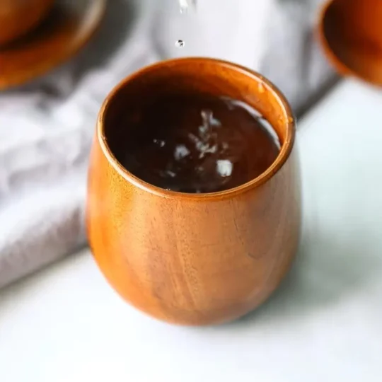 A close-up of a handcrafted wooden cup from The Artisan Sip, filled with a dark liquid, possibly tea or coffee, with another similar cup and saucer blurred in the background.