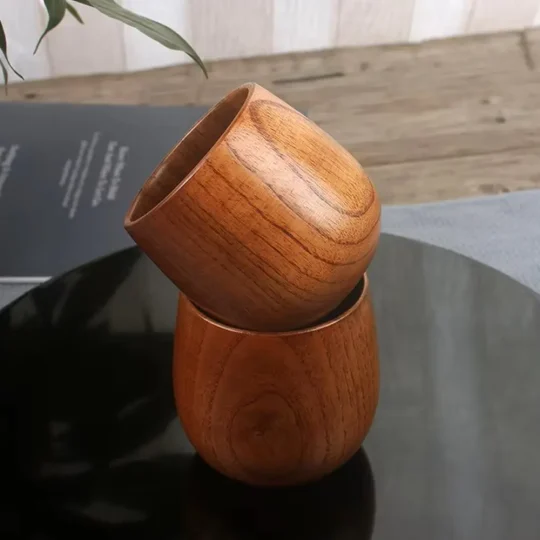 Two handcrafted wooden mugs from The Artisan Sip, one placed upside down on top of the other, showcasing the vertical grain pattern and warm brown color on a reflective black surface.