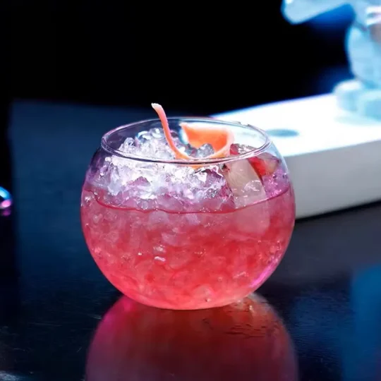 Close-up of a rounded glass with a pink beverage and ice, garnished with an orange peel twist, on a dark surface.