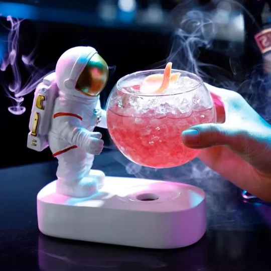 Hand holding a rounded glass with a playful astronaut design, filled with pink beverage and ice, emitting smoke, against a dark background.