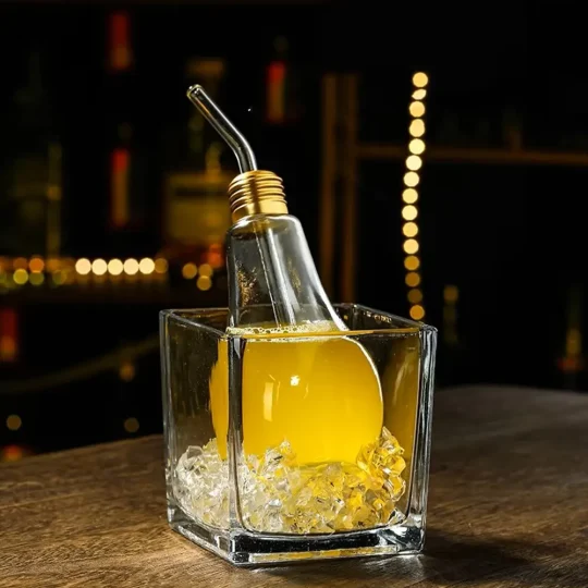 Clear glass with a yellow liquid and ice cubes, featuring a light bulb-shaped container inside filled with the same liquid and a metal straw, set against a dark background with blurred lights.