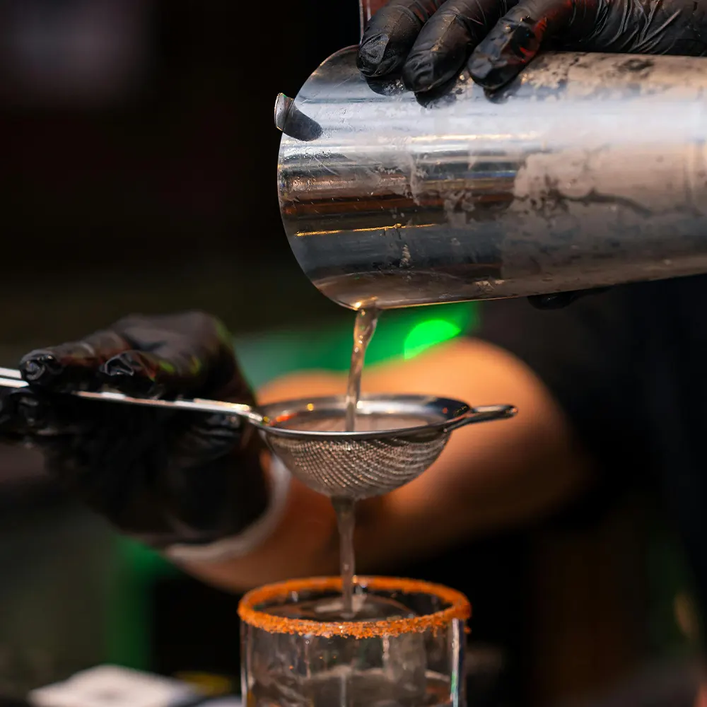 A close-up of a cocktail being poured through a strainer into a glass with an orange-rimmed edge, highlighting the versatility and elegance of premium drinkware for various beverages, from cocktails to seasonal drinks.