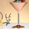 A copper-colored metallic martini glass made from food-grade stainless steel, filled with ice and garnished with a strawberry, with lemon slices and eyeglasses in the background.