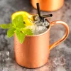 Close-up of a copper-plated stainless steel Moscow Mule mug with ice, mint sprig, lemon slice, and two black straws on a gray surface with another copper mug in the background.