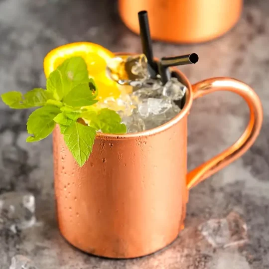 Close-up of a copper-plated stainless steel Moscow Mule mug with ice, mint sprig, lemon slice, and two black straws on a gray surface with another copper mug in the background.