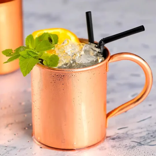 Copper-plated stainless steel Moscow Mule mug with ice, mint sprig, lemon slice, and two black straws on a wet surface.