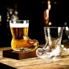 Two cowboy boot-shaped glasses on a wooden surface, one filled with amber beer and the other empty and tipped over, set against a dimly lit background with blurred bar elements.