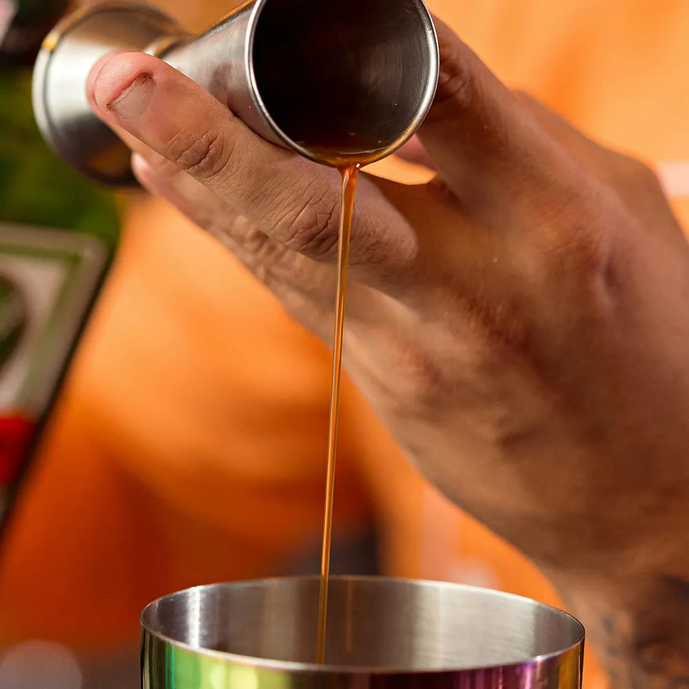 Hand holding a jigger, pouring liquor into a mixing tin, capturing the art of cocktail making.