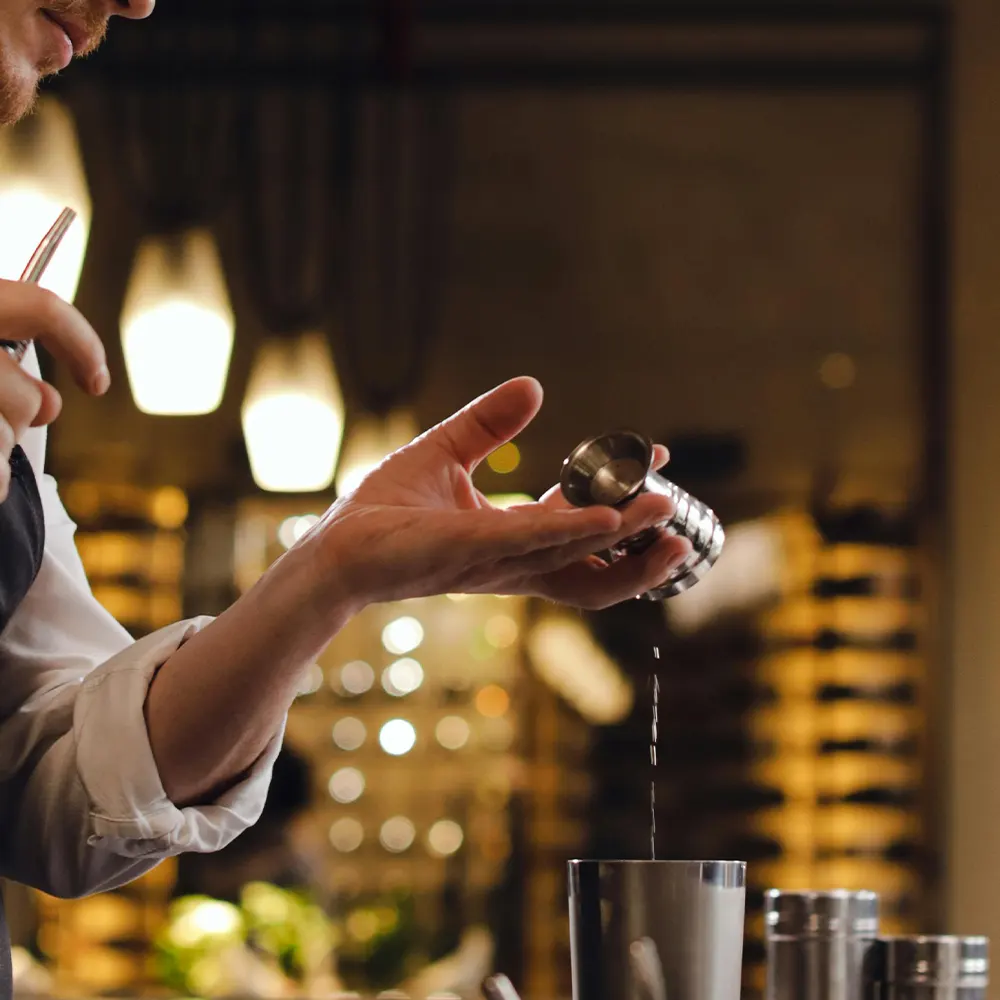 Close-up of hands pouring liquid into a metal jigger, capturing the essence of cocktail preparation in a warm, inviting setting.