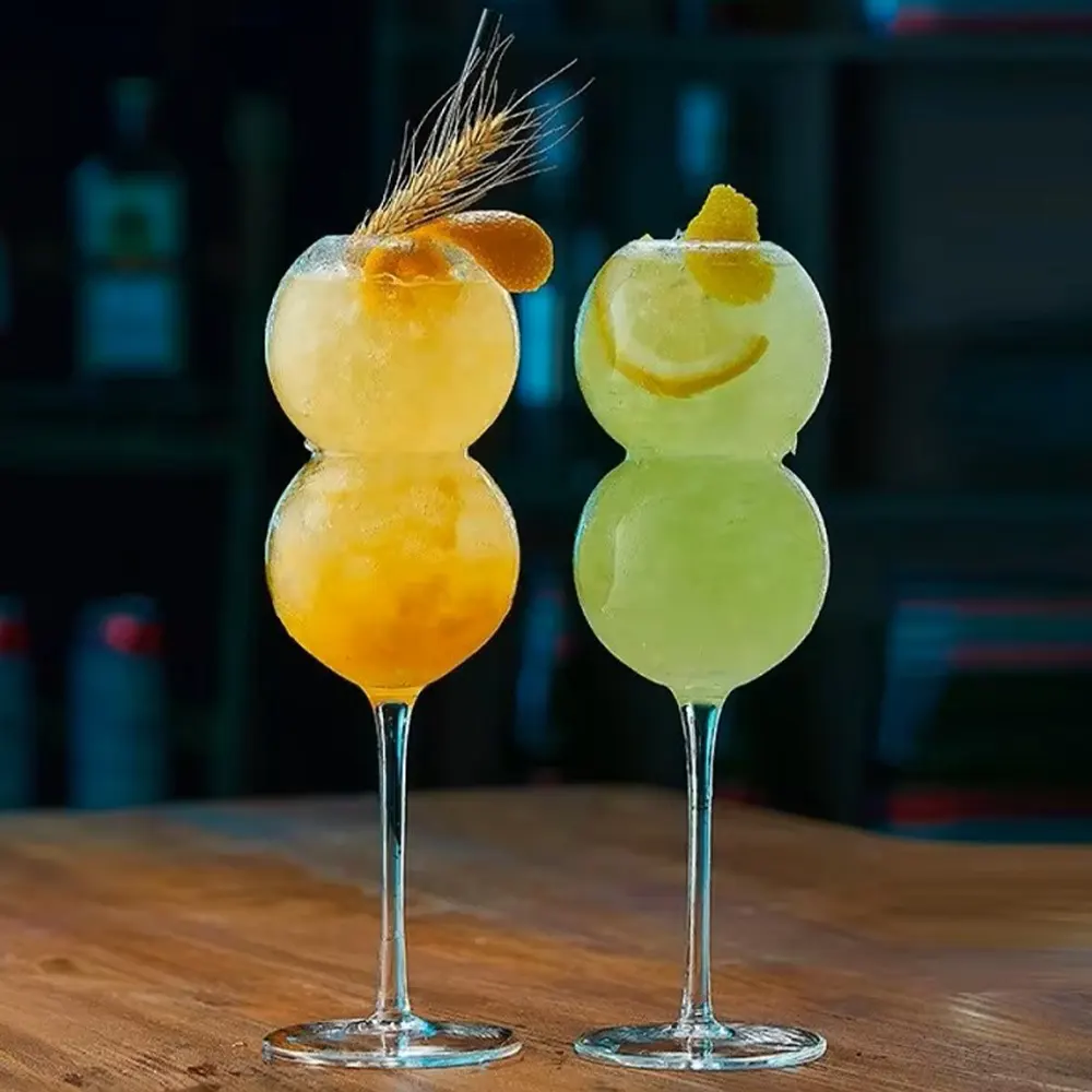 Set of two ‘The Double Bubble’ stemware glasses, each filled with a unique cocktail. The left glass contains an orange-hued drink garnished with wheat, while the right glass holds a green cocktail with a citrus peel garnish twisted into a smiley face. Both glasses are set against a blurred bar background.