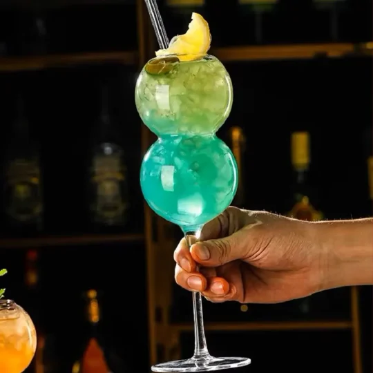 A hand holding ‘The Double Bubble’ stemware filled with a blue-green cocktail, featuring two spherical sections and a tall, slender stem. The top sphere is garnished with crushed ice and a slice of lemon, set against a dark background highlighting the vibrant drink.