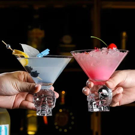 Two hands holding Skulltini Glasses with skull-shaped stems, one with a blue drink garnished with a lemon slice and umbrella, the other with a pink drink garnished with red fruit and green leaves.