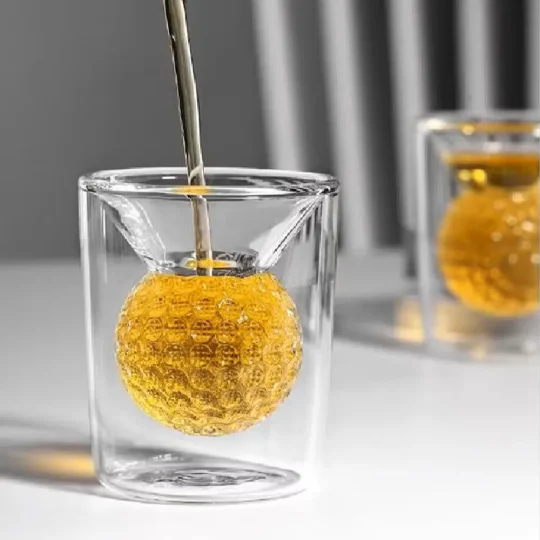 A Dual Illusion Tumbler with a clear outer square silhouette and a textured inner spherical section, filled with an amber-colored beverage being poured over the sphere, perfect for cocktails.