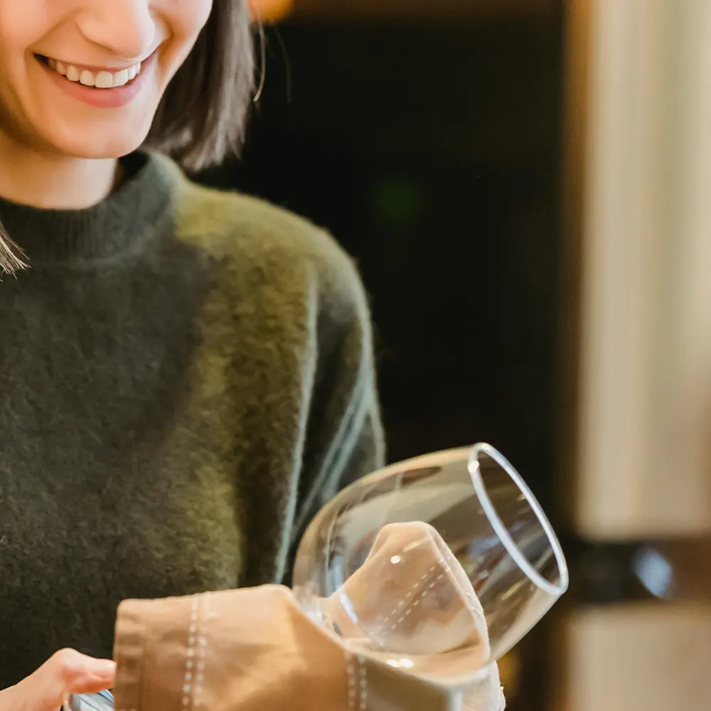 A person in a green sweater holding a clear glass, wiping it clean with a beige cloth, showcasing the ease of maintaining premium drinkware for enjoyable cocktail experiences.