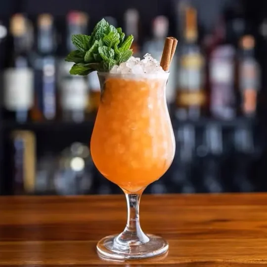 Curved cocktail glass with a refreshing cocktail, garnished with mint leaves and a cinnamon stick, showcasing elegant glassware against a blurred bar backdrop.