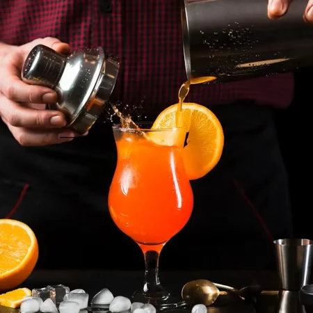 Close-up of a person pouring an orange beverage into a curved cocktail glass with an orange slice on the rim, set against a bar backdrop with ice cubes and bar tools.