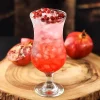 Curved cocktail glass filled with a red beverage, ice, and pomegranate seeds, placed on a wooden surface with pomegranate halves beside it.