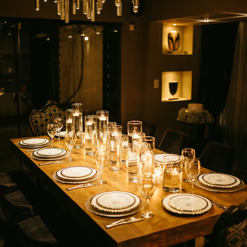 A dimly lit dining table set for a gathering, adorned with white plates, clear glasses, and candles, creating a sophisticated and inviting ambiance perfect for any occasion.