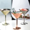 A collection of three metallic martini glasses in gold, silver, and copper finishes, made from food-grade stainless steel, placed on a reflective surface with a blurred indoor background.