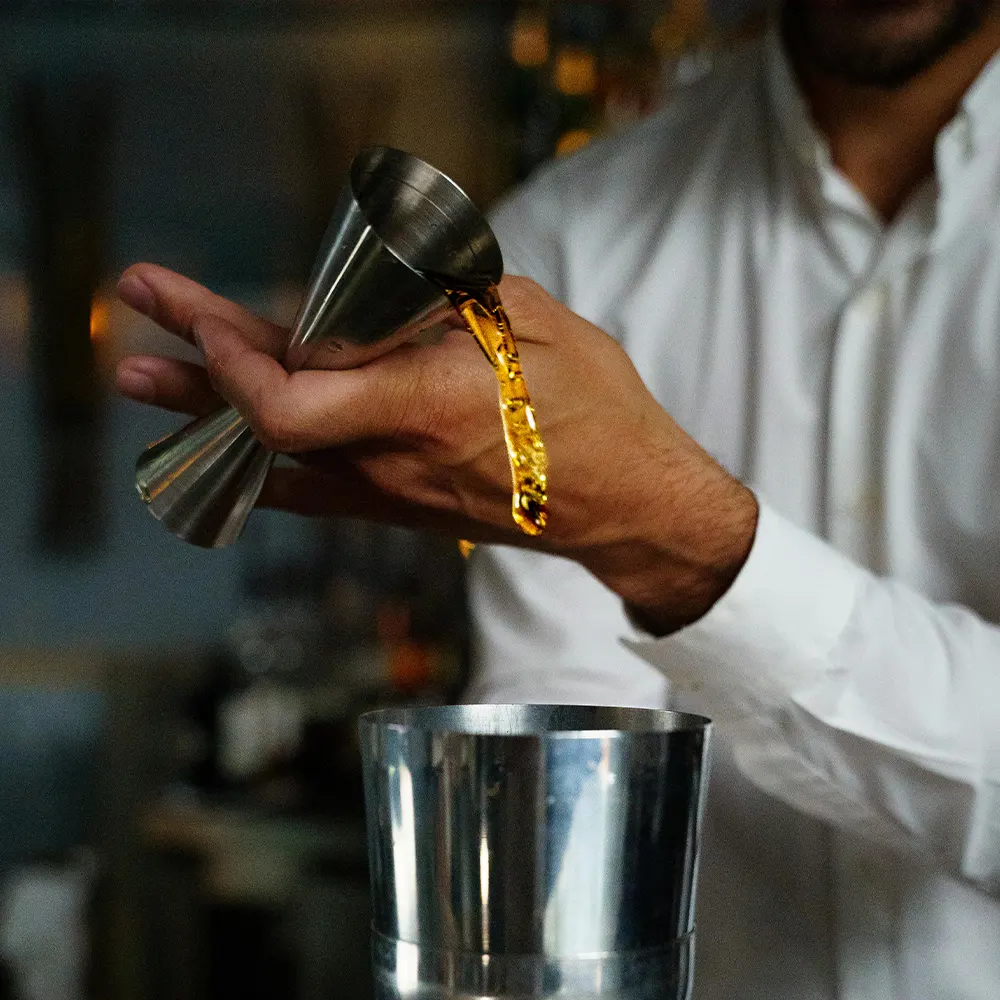 Hand pouring liquid from a small metallic jigger into a larger one, capturing the art of cocktail preparation.