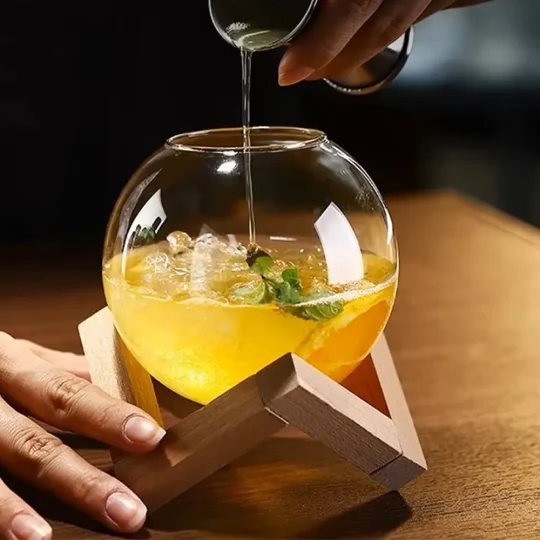 Hand pouring clear liquid into spherical 480 ml cocktail glass on geometric wooden stand with yellow-orange drink, ice cubes, and mint garnish