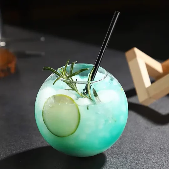 Spherical 480 ml cocktail glass with blue drink, lime slice, rosemary sprig, and black straw on wooden stand