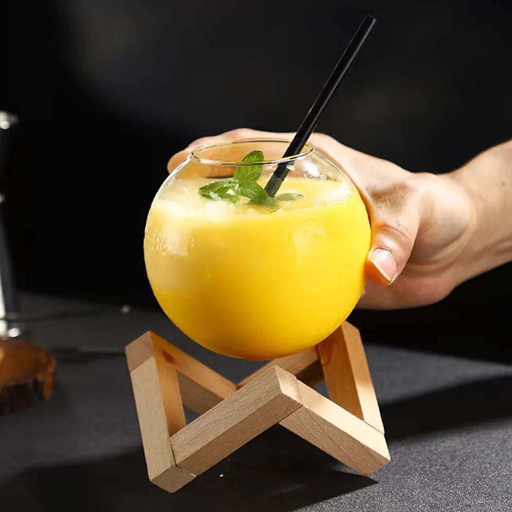 Hand Holding a Spherical 480 ml cocktail glass above an intricate wooden stand with orange drink, black straw, and mint garnish