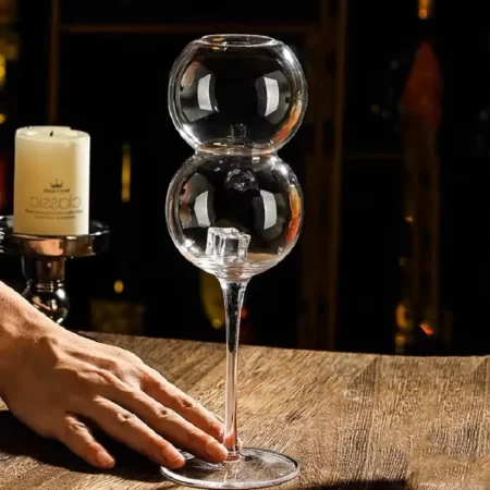 An empty ‘The Double Bubble’ stemware featuring two spherical chambers and a tall, slender stem, placed on a wooden surface with a hand resting beside it and a lit candle in the background.