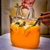 Hand holding the ‘Sip ‘n’ Carry’ handbag-shaped cocktail glass filled with a vibrant orange drink, garnished with citrus slices and mint leaves