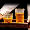 Two frosted silhouette glasses with a textured finish, filled with golden-brown liquid, placed on a wooden tray held by a person, against a blurred background.