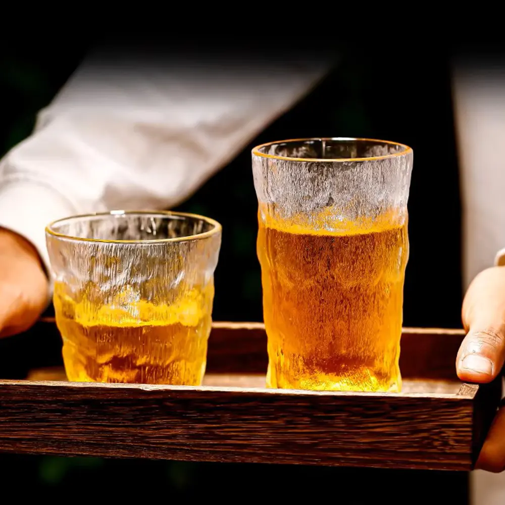 Two frosted silhouette glasses with a textured finish, filled with golden-brown liquid, placed on a wooden tray held by a person, against a blurred background.