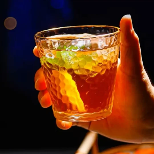 Close-up of a hand holding a Golden Cascade Tumbler with a hammered glass texture and gold rim, filled with a dark amber liquid and ice cubes, garnished with a lime wedge.