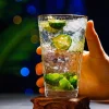 Close-up of a hand holding a Golden Cascade Tumbler with a hammered glass texture and gold rim, filled with a clear liquid and pieces of lime, against a festive background with blue and green bokeh lights.