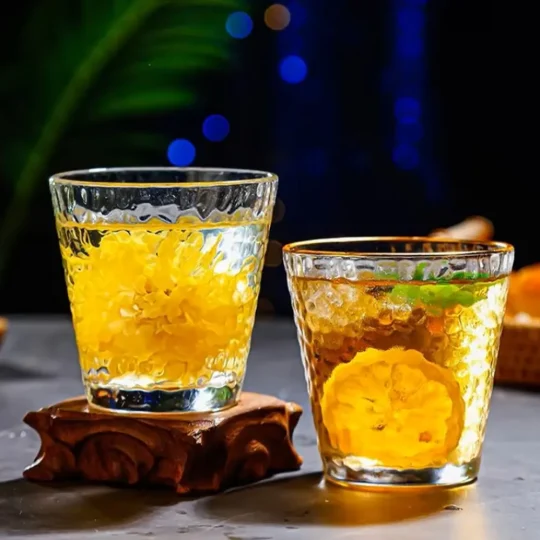 Two Golden Cascade Tumblers with a hammered glass texture and gold rim, filled with a yellow-orange beverage, ice cubes, and citrus slices, placed on a reflective surface with a blurred blue-lit background.