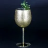 Close-up of a gold metallic wine glass with a smooth finish, filled with ice and garnished with mint leaves and a cherry, set against a dark background.