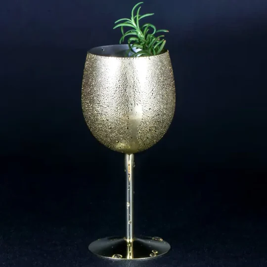 Close-up of a gold metallic wine glass with a smooth finish, filled with ice and garnished with mint leaves and a cherry, set against a dark background.