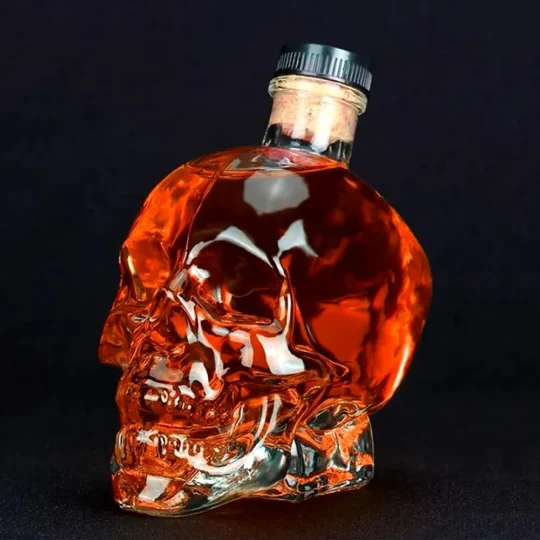 Clear glass skull-shaped Grim Reaper Bottle filled with dark amber spirit, featuring detailed facial features and a metallic cap, perfect for themed parties and collectors.