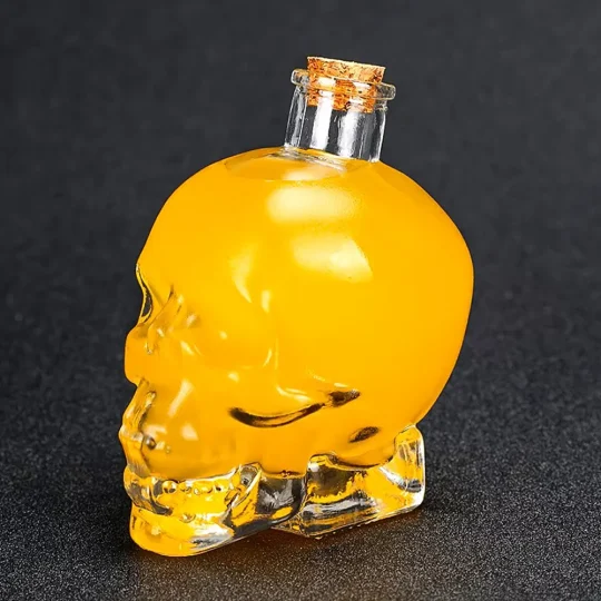 Small transparent glass skull-shaped Grim Reaper Bottle filled with yellow liquid, sealed with a cork stopper, against a dark grey background.