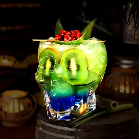 Skull-shaped Grim Reaper Glass filled with a layered green and blue cocktail, garnished with kiwi slices, red berries, and a green leaf, on a wooden surface.