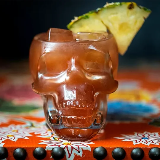 Skull-shaped Grim Reaper Glass filled with a brownish cocktail, garnished with a pineapple slice, set on an orange floral-patterned surface.