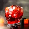 Skull-shaped Grim Reaper Glass filled with a red beverage, ice cubes, and pomegranate seeds, set against a warm, candle-lit background.